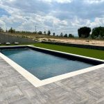 A modern rectangular swimming pool with clear water is surrounded by a stone patio and artificial turf