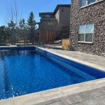 A rectangular blue pool in a backyard with stone patio, next to a stone house