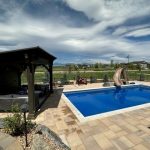Sunny backyard with a blue pool, slide, and stone patio. A shaded pergola with seating is on the left